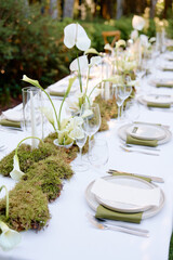A chic outdoor dining scene featuring a long white table, moss runners, tall calla lilies, and refined place settings with napkins, plates, and crystal glassware, creating a serene,