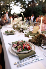 A festive outdoor dinner scene featuring a long white table, candlelight, greenery, and a plated salad with meat slices. Guests mingle in the background, creating a warm, celebratory atmosphere.