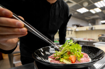 Chef plating gourmet beef carpaccio with salad and strawberries in professional kitchen. Culinary precision, fine dining presentation, stainless steel background.