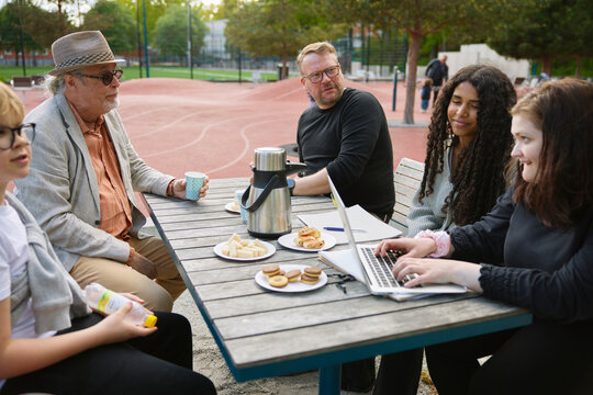 Family friends spending leisure time eating snacks and having tea while sitting at bench in park