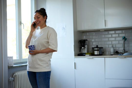 Smiling woman talking on smart phone while standing in kitchen at home