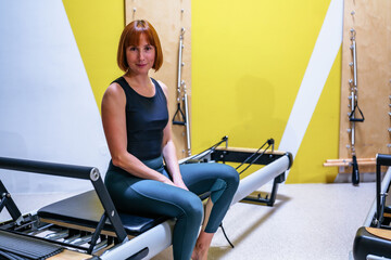 Woman smiling on a pilates reformer, seated in activewear during a modern studio workout, pausing...