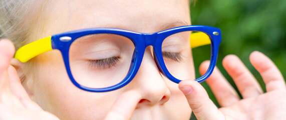 Closeup child’s face wearing blue and yellow glasses, moment of quiet concentration, focused gaze, bright background, childhood vision care, stylish eyewear for kids, playful and thoughtful expression