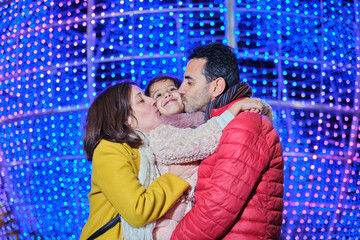 Parents kissing daughter under colorful Christmas lights at night