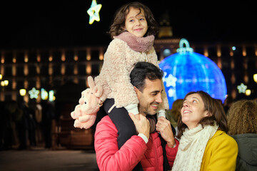 Father carrying daughter on shoulders in festive Christmas square at night