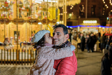 Father smiling as daughter kisses him with carousel lights at night