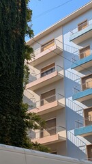 Vertical architectural photo showing a corner of a Spanish building covered with climbing greenery and two colorful houses in yellow and blue tones. Bright sunlight and soft shadows create a clean