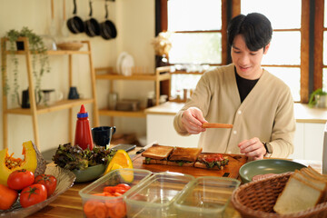Man Making Sandwiches at bright and minimalist Counter with Fresh Greens, ham. Asian man cooking healthy foods in cozy kitchen in morning at home.