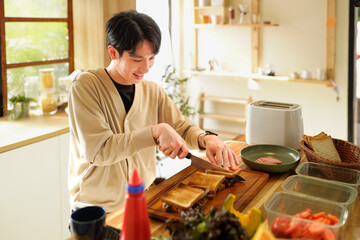 A man is preparing a sandwich on a wooden cutting board. The kitchen is well-stocked with various food items