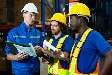 Group of three diverse warehouse workers collaborating and discussing logistics planning, holding clipboard and rolled paper documents, wearing safety helmets inside industrial warehouse