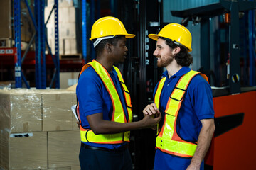Warehouse scene with two diverse male workers shaking hands wearing helmets and safety vests smiling warmly demonstrating teamwork success and mutual respect inside industrial storage area