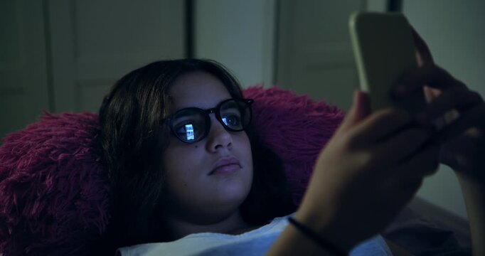 A teenager in bed, wearing glasses, looks at her mobile phone, which reflects her as she quickly scrolls through the pages on her phone. In the darkness of the night, in her room. Medium shot.