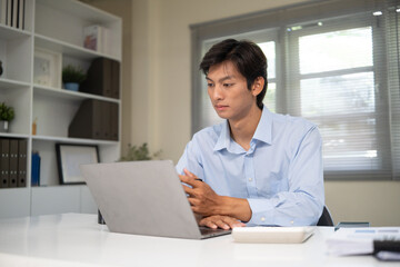 Fototapeta premium Young businessman working on laptop at modern office desk, surrounded by financial documents.