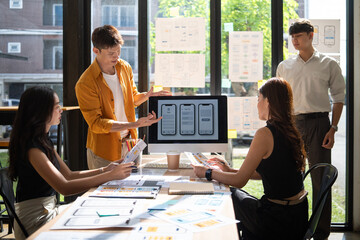 Group of young professionals collaborating on a user interface project, reviewing wireframes and mobile app mockups on computer screen.