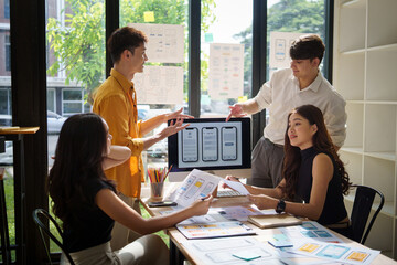 Group of young professionals collaborating on a user interface project, reviewing wireframes and mobile app mockups on computer screen.