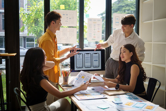 Group of young professionals collaborating on a user interface project, reviewing wireframes and mobile app mockups on computer screen. - Powered by Adobe