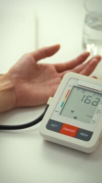 A middle-aged woman measures her blood pressure in a bright, room-like room. She uses a special, high-tech electronic device to measure blood pressure at home