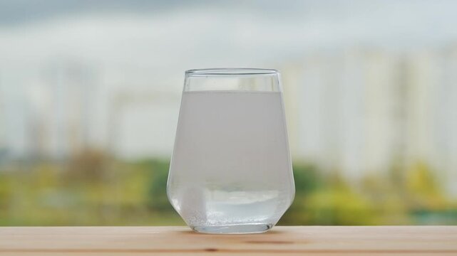 Vitamin tablet slowly dissolves in water with fizz and clouding, creating a healthy drink in clear glass against soft autumn cityscape.