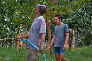 A father waters a young mango tree with a hose while carefully examining the leaves and growth, as his son stands nearby observing plants across their farmland.