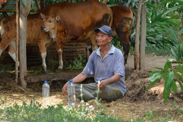 A farmer rests on the ground beside dried cow dung, looking thoughtfully at his farmland with a plastic water bottle placed in front of him.