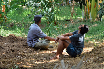 Sitting on the soil in front of the cow shed, a farmer and his son calmly observe the plants and trees growing in their peaceful farmland.