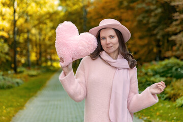 Female model in pastel pink hat and scarf lifts a plush heart while standing on a tree-lined park...