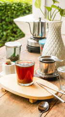 A tranquil scene of Vietnamese drip coffee or tea on a rustic wooden table with a white plant vase and lush green outdoor background