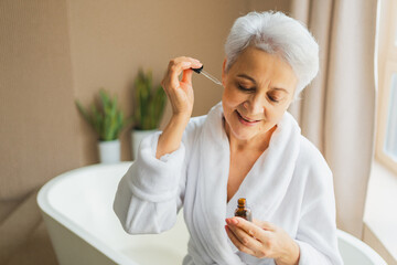 Woman applying natural drops cosmetics oil to face skin. Mature adult old woman applying nourishing anti age serum with pipette doing daily beauty routine. Senior woman gets kin scare facial treatment