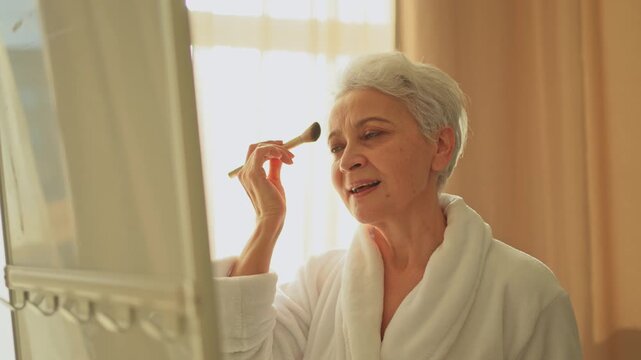Mature adult old woman with smooth soft perfect skin applying rouge with brush on cheek looking at mirror doing daily beauty make up routine. Senior woman putting blush on her face. Anti age makeup