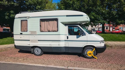 Wide view of campervan with yellow wheel clamp parked on brick sidewalk near houses © Kira_Yan