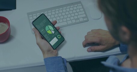 Holding smartphone displaying data charts, worker wearing blue shirt at desk with keyboard, red mug