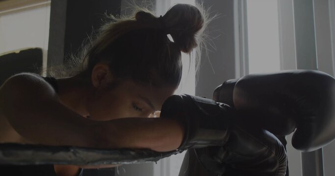 Leaning boxer resting on ring ropes inside boxing gym with gloves, sleeveless top and window blinds