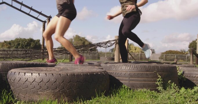 Leaping workout partners negotiating grassy field obstacle course with tire hurdles and metal frame