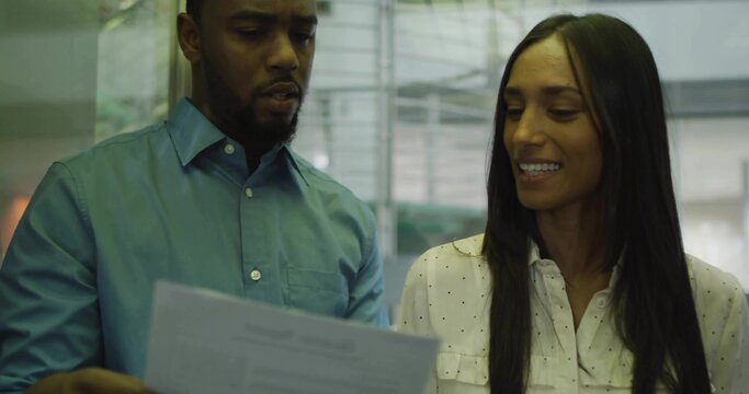 Standing colleagues wearing teal shirt and dotted blouse reading printed document in office lobby