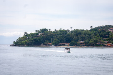 Obraz premium A small motorboat cruises across calm coastal waters, with lush tropical vegetation, and waterfront houses lining the shoreline of the island on an overcast day