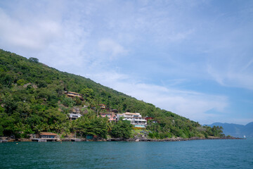 Hillside homes with terracotta rooftops line the mountainside along the Atlantic coast, surrounded by lush greenery under a bright blue sky in Ilhabela, Brazil