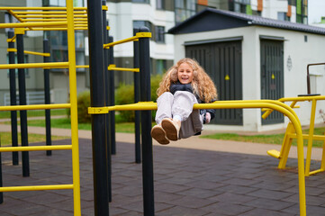Obraz premium joyful girl playing on monkey bars in urban playground, wearing casual attire. bright yellow play equipment contrasts with surrounding modern architecture, capturing childhood fun
