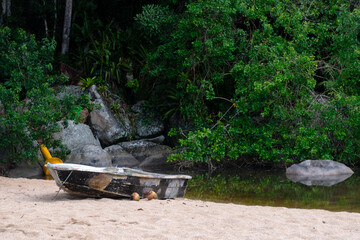 An old weathered boat rests on a sandy shore in Ilhabela, Brazil, beside lush green forest and calm water, evoking a rustic tropical beach scene