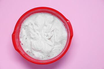 Plastic basin with clothes, water and foam on pink background, top view