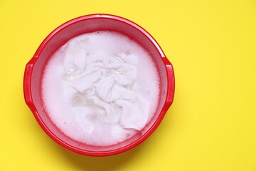Plastic basin with clothes, water and foam on yellow background, top view. Space for text