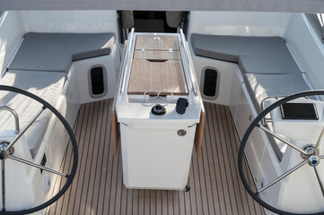 Sailboat with dual steering wheels, comfortable cabins with grey cushions, and wooden decking. Central table with compass and modern design, perfect for a comfortable and luxurious sailing experience.