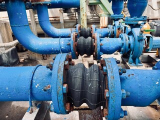 Industrial blue water pipes with rusted flanges connected by a flexible rubber expansion joint to absorb vibration and movement in the pipeline system.