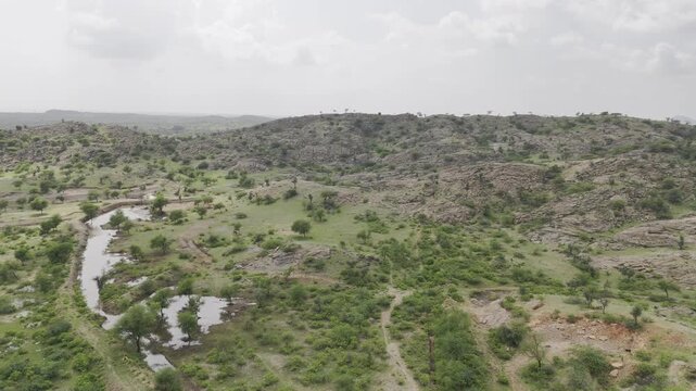 Aerial shot of rocky hills near Masuda, Rajasthan, showing green Aravalli vegetation, small ponds, and water bodies under a cloudy monsoon sky, creating a serene rural landscape.