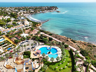 Drone shot over Cabo Roig, Costa Blanca, view of swimming pool near rocky seashore, green lawns, palm trees, villas, and scenic bay with turquoise Mediterranean waters and sandy beach