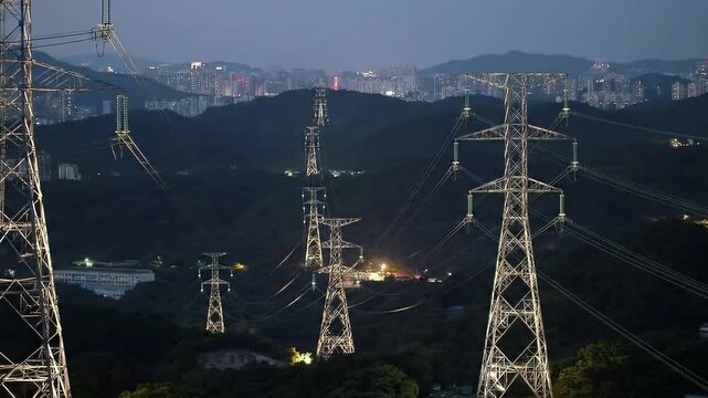 A network of towering power pylons stretches across a shadowy mountainous landscape at dusk, forming a symbol of industrial energy. 