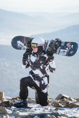 A snowboarder sits on his knees at the top of a mountain, holding a snowboard on his shoulders.