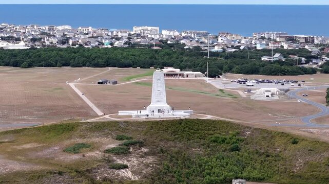 An expansive aerial shot shows a circular road loop around a grassy hill topped by a White Wright Brothers monument, with distant Kill Devil Hills and Outer Banks Coastline under a clear blue sky.