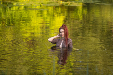 Female figure with red hair is immersed in calm water, surrounded by vibrant greenery, evoking a...