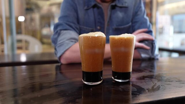 Two glasses of dark stout beer with a cascading nitro effect on a wooden table. A man in the background is waiting for the tasting