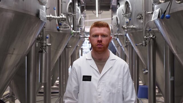 Young handsome brewmaster with red hair wearing a white lab coat, standing between rows of steel fermentation tanks in a beer factory